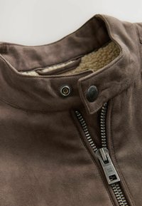 Chaqueta de ante marrón con un alto cuello, cierre de cremallera y forro interior de felpa. Los elementos metálicos destacados incluyen botones a presión y una cremallera plateada.