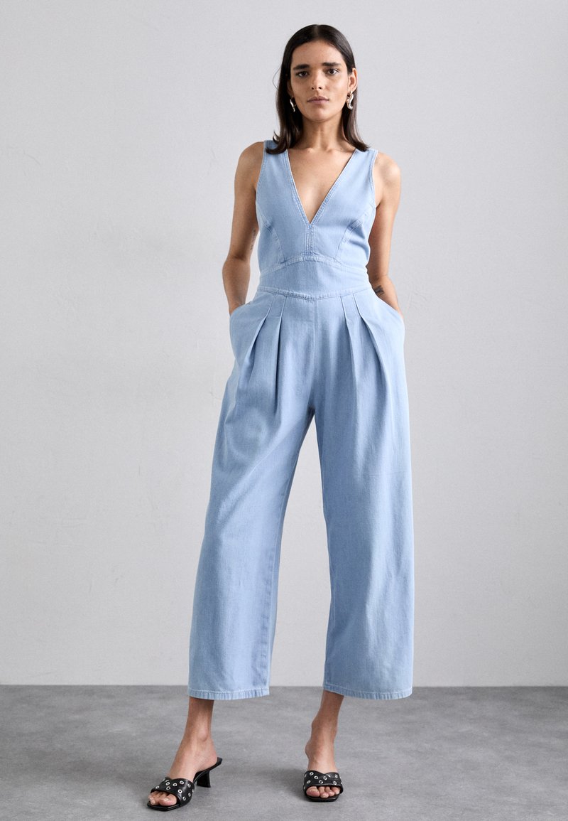 Jumpsuit en denim bleu clair avec un décolleté en V profond, taille plissée et jambes larges. Comprend des poches et est associé à des mules noires à talons.