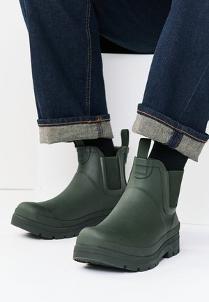 Sötétzöld vízálló bokacsizma és felhajtott sötétkék farmernadrágot viselő személy, fehér felületen ülve.