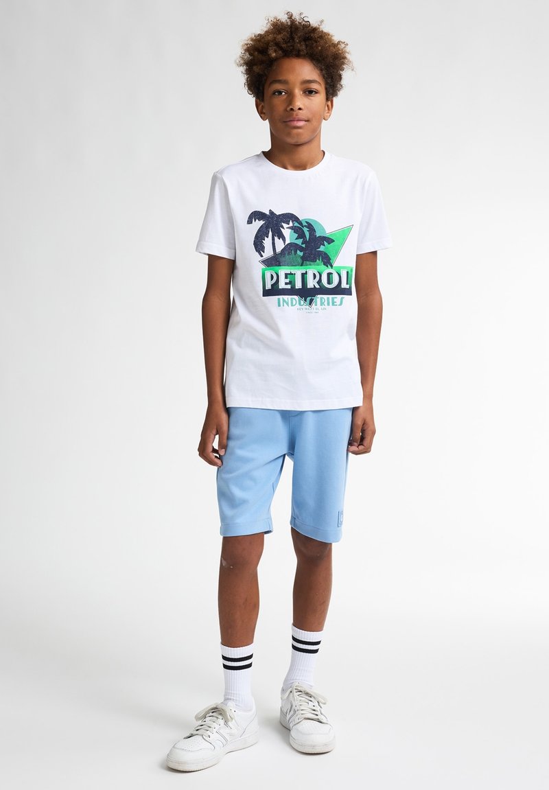 Witte katoenen t-shirt met groen en marineblauw grafisch ontwerp. Lichtblauwe shorts, kniehoog met opgerolde mouwen. Witte sneakers met zwarte strepen.