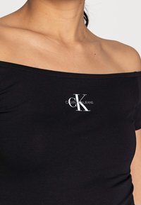 Czarny t-shirt z dekoltem typu off-the-shoulder wykonany z miękkiego materiału, z białym logo "CK CALVIN KLEIN JEANS" na przodzie.
