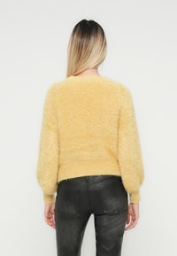 Vrouw met lang ombre-haar, gekleed in een fuzzy gele trui en zwarte neplederen broek, staande met haar rug naar de camera.