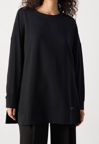 Czarny sweter z długimi rękawami, okrągłym dekoltem i luźnym krojem. Miękka faktura z bocznym rozporkiem i subtelnym logo w pobliżu dolnej krawędzi.