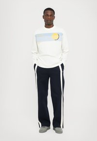 Cămașă albă cu mâneci lungi, având un design grafic albastru și galben. Pantaloni bleumarin cu dungi laterale albe. Modelul stă relaxat.