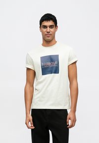 Kremowa koszulka z krótkim rękawem z prostokątnym niebieskim nadrukiem i czerwonym napisem "BARBOUR INTERNATIONAL". Gładka bawełniana tekstura.