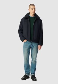 Giubbotto bomber blu navy con colletto in pelliccia sintetica nera, indossato sopra un maglione verde, abbinato a jeans azzurri e scarpe nere.