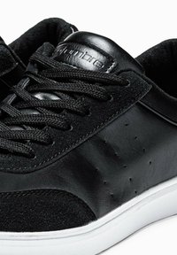 Sneaker nera con una combinazione di pelle scamosciata liscia e testurizzata, lacci rotondi, perforazioni sottili e suola in gomma bianca.