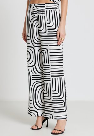 Frau trägt weiße Hose mit hoher Taille und weitem Bein mit schwarzen geometrischen Linienmustern und schwarzen, hochhackigen Sandalen mit Riemchen.