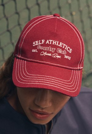 Piros baseballsapka fehér hímzett felirattal: „Self Athletics Country Club est. Sports Dept. 2023.” Kontrasztos varrással.