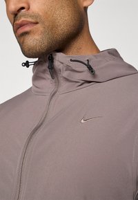 Nike jacket in light grey fabric, featuring a zip-up front, drawstring hood, and subtle logo accent. Smooth texture with elastic details.