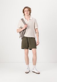 Svijetlo bež pleteni polo s kragnom, maslinasto zeleni šortsevi, sivi ranac, bijele tenisice s smeškim detaljima. Jednostavan, ležeran i dobro krojen outfit.