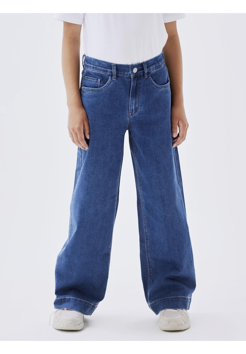 Personne portant un jean bleu large, un t-shirt blanc et des baskets blanches, debout les mains détendues le long du corps sur un fond clair.