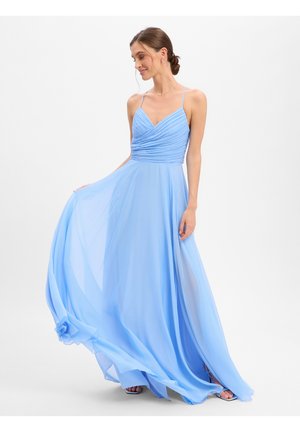 Femme dans une robe fluide bleu clair sans manches avec de fines bretelles, tenant la jupe en regardant vers le bas et souriant doucement.