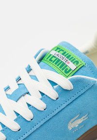 Modré semišové tenisky s bílými tkaničkami. Obsahují zeleno-bílou etiketu na jazyku s nápisy "LACOSTE" a "TENNIS." Zahrnují detail loga na boku.