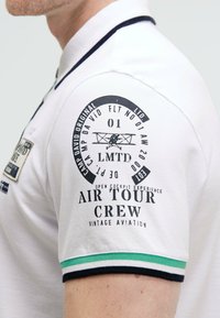 Weißes Poloshirt mit einem schwarzen Grafikdesign am Ärmel, das flugzeugbezogenen Text enthält. Akzentuiert mit marineblauen und grünen Streifen am Kragen.