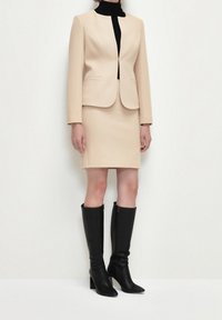 Beige räätälöity bleiseri, jossa on rakenteelliset olkapäät, yhdistettynä vastaavaan kynähameeseen, jota täydentävät mustat polvimittaiset korkokengät.
