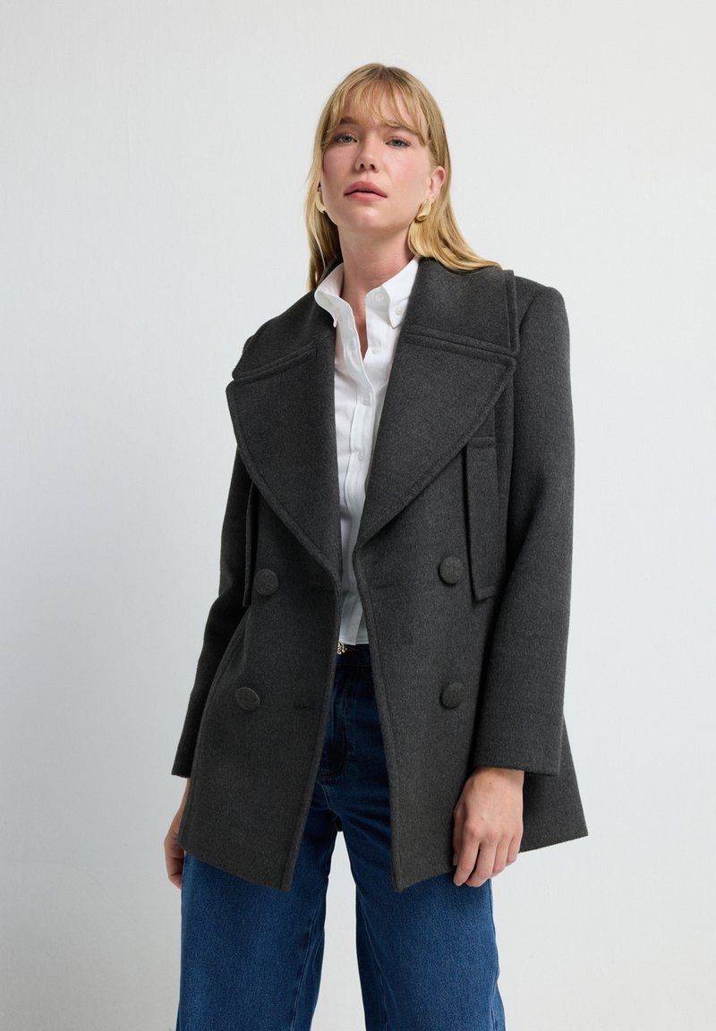 Abrigo de lana gris tipo peacoat con diseño de doble botonadura, grandes solapas y bolsillos laterales, combinado con una camisa blanca y jeans azules.