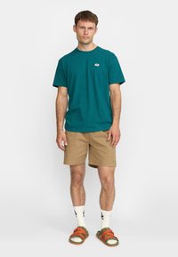 Camiseta de algodón verde azulado con mangas cortas y un pequeño logo, combinada con pantalones cortos beige, calcetas blancas y sandalias con tiras naranjas.