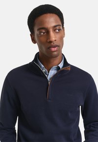 Pólder azul marino con cuello alto, que cuenta con una cremallera y un acento de cuero marrón. Tejido texturizado, superpuesto sobre una camisa de cuello azul claro.