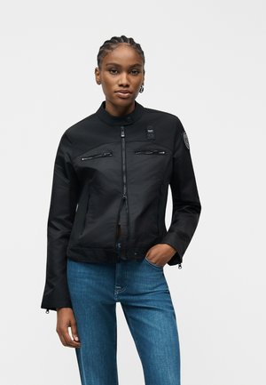 Γυναίκα με πλεξουδάκια, φορώντας μαύρο μπουφάν με φερμουάρ και μπλε τζιν, στέκεται με το ένα χέρι στην τσέπη, μπροστά σε απλό φόντο.