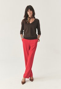 Donna in piedi con le mani in tasca, veste un maglione marrone a scollo a V, pantaloni rossi, tacchi marroni, braccialetto marrone e una sciarpa a stampa leopardata.