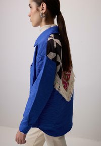 Giacca blu trapuntata con tessuto stampato sul retro, con dettagli in frange. Il modello indossa un dolcevita e orecchini a cerchio in argento.