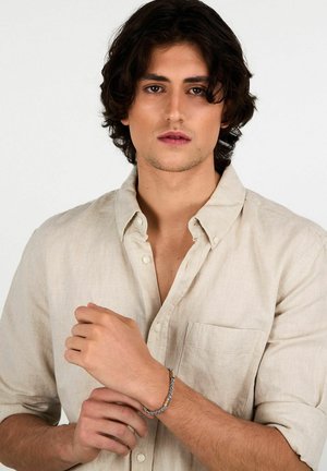 Bracciale intrecciato in argento con accenti gialli indossato al polso di una persona con una camicia beige chiaro a bottoni, con uno sfondo neutro.