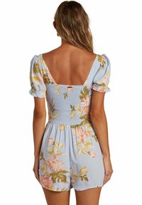 Blåblommig jumpsuit med korta puffärmar, smockad rygg och djup V-ringning. Den har stora rosa och gula blommönster överallt.