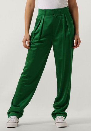 Osoba ubrana w błyszczące, zielone spodnie z wysokim stanem i plisami, zestawione z białymi trampkami i białym, bezrękawnym topem, stojąca na jednolitym tle.