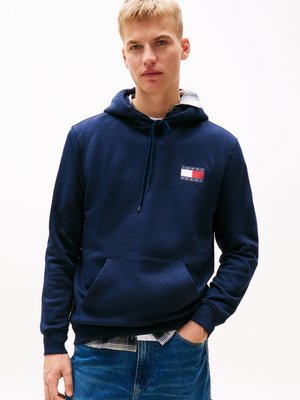 Marineblauwe hoodie van katoen, met een voorste kangoeroezak en een logo-label in rood en wit op de borst, gedragen over een geruit overhemd.