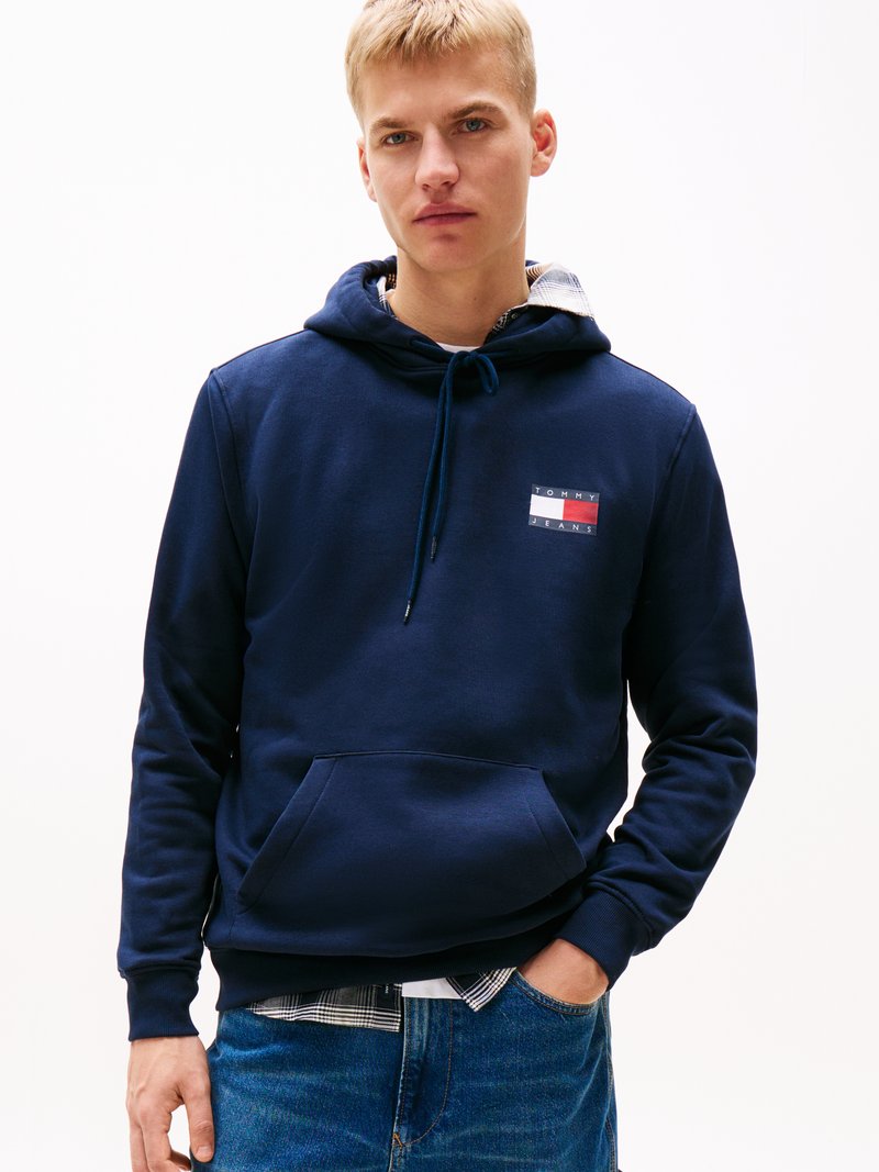 Un hanorac de culoare bleumarin, fabricat din bumbac, având o buzunar frontal tip kangaroo și o pată cu logo alb și roșu pe piept, purtat peste o cămașă în carouri.