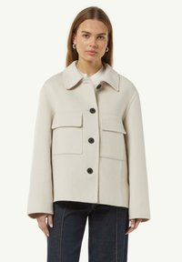 Lichtbeige jas van zacht materiaal, met een kraag, vier voorzakken en zwarte knopen. Vierkante snit met lange mouwen.