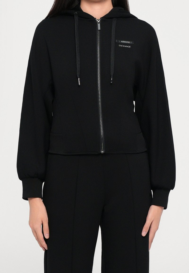 Femme portant un sweat à capuche noir zippé avec poches devant et manches longues, assorti à un pantalon noir.