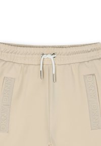Beige joggingbroek met elastische tailleband, een trekkoordsluiting en tonale DKNY-branding aan de zijkanten. Glad, lichtgewicht materiaal.