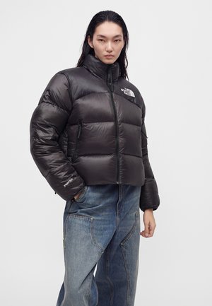 Млада жена, облечена в черно пухено яке с логото на The North Face и сини широки дънки, стои с ръце частично в джобовете.