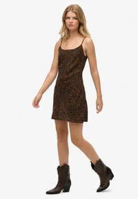 Vestido marrón con estampado de leopardo, con tirantes finos, corte hasta la rodilla y un ajuste holgado. Combinado con botas de tobillo de estilo cowboy marrón.
