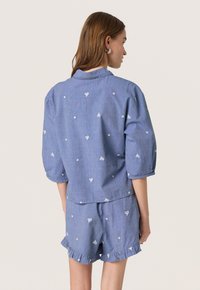 Camicia in chambray blu con ricami floreali bianchi, maniche a sbuffo e orlo curvo, abbinata a pantaloni corti coordinati con bordo arricciato.