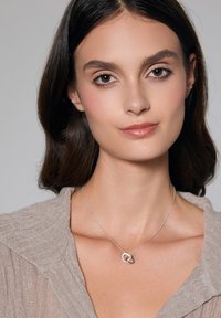 Srebrna naszyjnik z przeplatającymi się okrągłymi ogniwami, noszony przez modelkę w jasnobeżowej bluzce, na neutralnym tle.