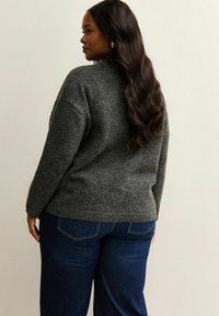 Femme avec de longs cheveux noirs portant un pull gris foncé et un jean bleu, légèrement tournée sur le côté devant un fond clair uni.