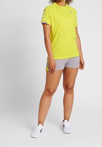 Ljust gul kortärmad träningströja med grå detaljer, matchad med grå shorts som har gula ränder. Modellen har på sig vita sneakers.