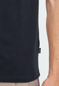 Widok z boku na osobę w czarnej koszulce z małą czarną metką z napisem "Casual Friday" w pobliżu brzegu i beżowych spodniach.