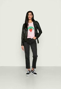 Jaqueta de motociclista de couro preto, t-shirt gráfica rosa com texto verde e morango vermelho, jeans pretos e ténis pretos com solas brancas.