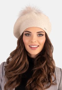 Beret en tricot beige avec un pompon duveteux sur le dessus. Le tissu semble doux avec une finition texturée. Détail du logo visible sur le côté.