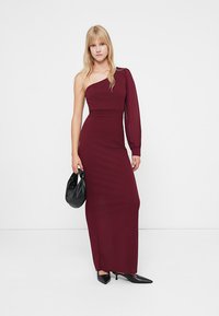 Βουργουνδί δροσερό φόρεμα μίας ώμου με εφαρμοστή σιλουέτα· ένα μακρύ μανίκι από διαφανές ύφασμα· συνδυασμένο με μαύρη τσάντα και στιλέτο παπούτσια.