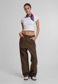 Weißes, gekürztes T-Shirt kombiniert mit locker sitzenden, dunkelbraunen Hosen. Das Outfit wird durch einen bunten Schal um den Hals und weiße Sneakers ergänzt.