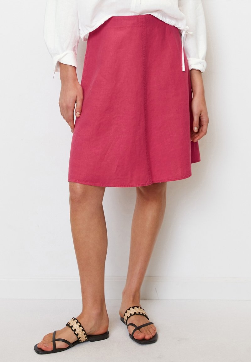 Falda A-line de lino rosa, de longitud hasta la rodilla, con una textura suave. Combinada con sandalias planas negras que tienen una correa con un estampado.