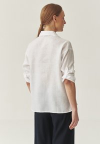White linen shirt with rolled-up sleeves, a collar, and a relaxed fit. Soft texture and lightweight fabric, shown from the back.