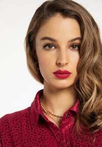 Rot gepunktetes Button-up-Hemd mit Kragen. Schmuck umfasst eine schichtweise goldene Kettenhalskette. Lange wellige braune Haare.