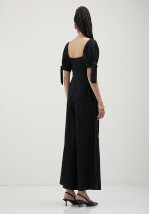 Frau mit geflochtenem Haar, die einen schwarzen Jumpsuit mit Puffärmeln und Schleifendetails, schwarze Slingback-High Heels und goldene Armbänder trägt, stehend und mit dem Rücken zur Kamera.