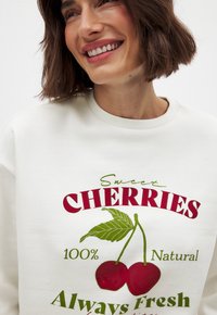 Hellbeige Sweatshirt mit einem Kirsch-Grafikdesign in Rot und Grün sowie dem Schriftzug "Süße Kirschen 100% Natürlich Immer Frisch", der gut sichtbar ist.
